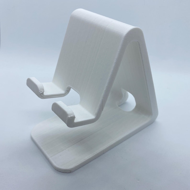 Peut inclure: Support de t&eacute;l&eacute;phone en plastique blanc avec un design simple. Le support a deux ergots pour maintenir un t&eacute;l&eacute;phone en position verticale.