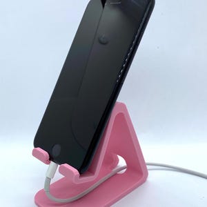 Peut inclure: Un support de t&eacute;l&eacute;phone rose en plastique avec un smartphone noir pos&eacute; dessus. Le t&eacute;l&eacute;phone est branch&eacute; &agrave; un c&acirc;ble de charge blanc.