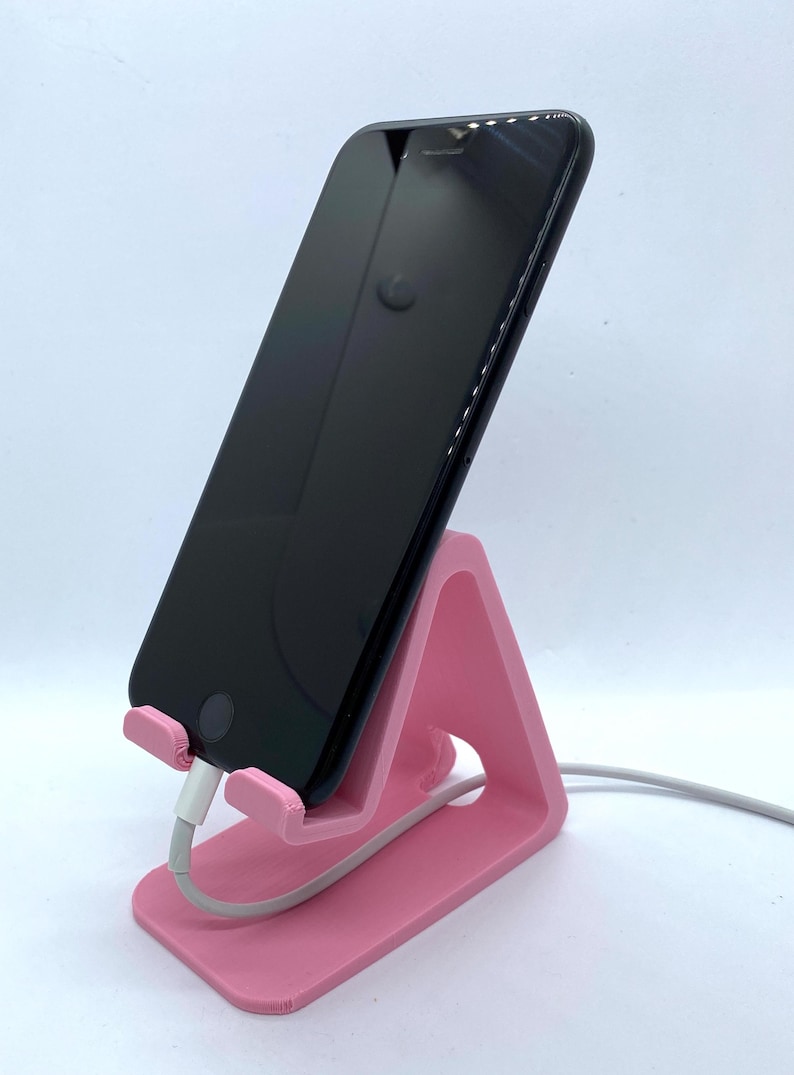 Peut inclure: Un support de t&eacute;l&eacute;phone rose en plastique avec un smartphone noir pos&eacute; dessus. Le t&eacute;l&eacute;phone est branch&eacute; &agrave; un c&acirc;ble de charge blanc.