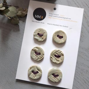 May include: A set of six round, light green ceramic buttons with textured surfaces and dark purple thread accents, attached to a white display card labelled with the text MMK Miss Muffet's Knits, Elisha Orchard, and Made In Mayfield, New Zealand.