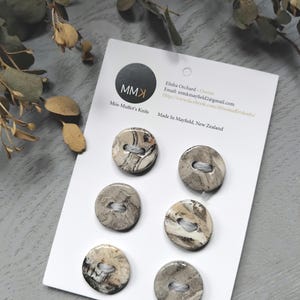 Op de afbeelding: Zes ronde, gemarmerde knopen in grijstinten en crème worden gepresenteerd op een witte kaart. De kaart bevat de tekst "Miss Muffet's Knits" en contactgegevens. De knopen hebben een diameter van ongeveer 2,5 cm.