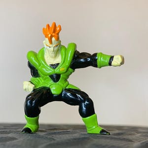 May include: A small figurine of a character in a fighting stance. The figure has orange hair, a pale face, and is wearing a green and black outfit. The figure is posed with one arm extended and the other bent, as if ready to fight. The figurine is on a textured surface.