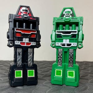 May include: Two plastic robot action figures standing side by side. One figure is primarily black with red and silver accents, while the other is green with silver accents. Both robots feature a blocky, mechanical design with square details on the feet.