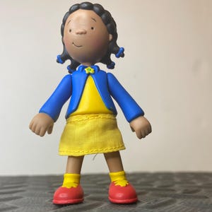 以下が含まれることがあります： 黒い髪をツインテールに結った、褐色の肌のキャラクターのプラスチック製フィギュア。青いジャケット、黄色いシャツ、黄色いスカート、黄色い靴下、赤い靴を着用している。襟元には小さな黄色い花の装飾が施されている。