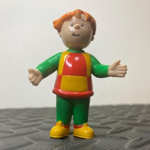 以下が含まれることがあります： オレンジ色の髪、そばかす、笑顔が特徴的なキャラクターの小さなプラスチック製フィギュア。緑色の長袖シャツに赤と黄色のベスト、緑色のズボン、黄色の靴を着用しており、両腕を横に広げたポーズで立っている様子が表現されています。