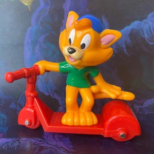 May include: A plastic figurine of an orange cat with a blue cap and green shirt riding a red three-wheeled scooter. The cat has large white eyes, a black nose, and a pink mouth. The scooter features a red handlebar and red wheels.