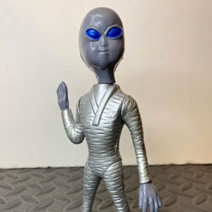 May include: A gray alien figurine with a smooth, rounded head and large, vibrant blue oval eyes. The figure wears a textured, silver-colored suit that covers the body from the neck down to the feet, with one arm raised in a greeting gesture.