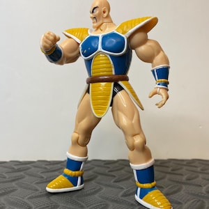 May include: A bald, muscular action figure with a tan complexion, wearing blue and yellow segmented armor with white trim. The figure features a brown belt, blue and white wrist guards, and blue and yellow boots, standing in a dynamic pose.