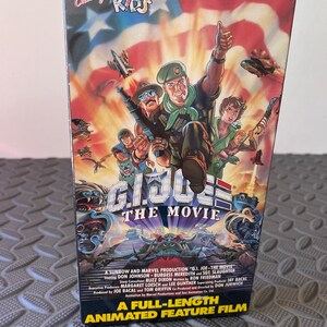 Puede incluir: Una carátula de cinta VHS para la película animada "G.I. Joe: The Movie". La carátula presenta una ilustración colorida de los personajes de G.I. Joe, incluidos Duke, Snake Eyes y Scarlett, sobre un fondo de bandera estadounidense y explosiones. El texto "A Full-Length Animated Feature Film" está impreso en la parte inferior de la carátula.