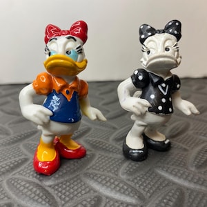 May include: Two small plastic figurines of a cartoon duck character. The left figure wears an orange top, blue shirt, and red shoes with a red bow. The right figure is painted in black and white, featuring a black polka-dot dress and bow.