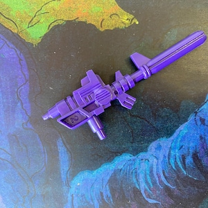 May include: A purple toy gun with a detailed design, featuring a barrel, scope, and handle. The toy gun is made of plastic and has the number "2" printed on the side. The background is a colorful abstract painting with shades of blue, green, and black.