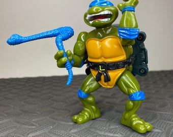 Pode incluir: Figura de ação de Leonardo das Tartarugas Ninja. A figura verde tem uma máscara azul, um peito amarelo e um cinto preto. Ele segura uma arma azul e está em uma pose dinâmica. Mede aproximadamente 15 cm de altura.