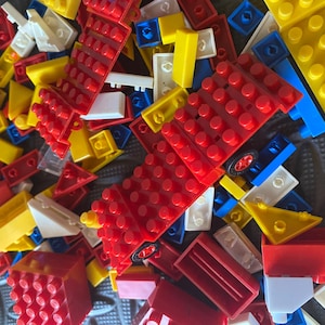 May include: A pile of colourful plastic building bricks in red, yellow, blue, and white. The bricks are of various shapes and sizes, including rectangular, square, and triangular pieces. Some bricks have circular studs.