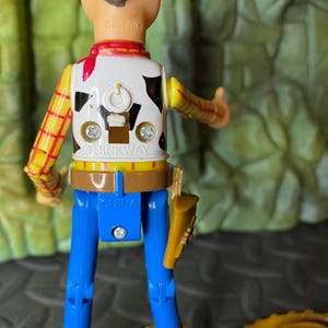May include: A Woody action figure from the Toy Story franchise. The figure has a white vest with a black cowhide pattern, a yellow and red plaid shirt, blue pants, and a brown belt with a holster. The figure is marked "THINKWAY" and "CHINA".