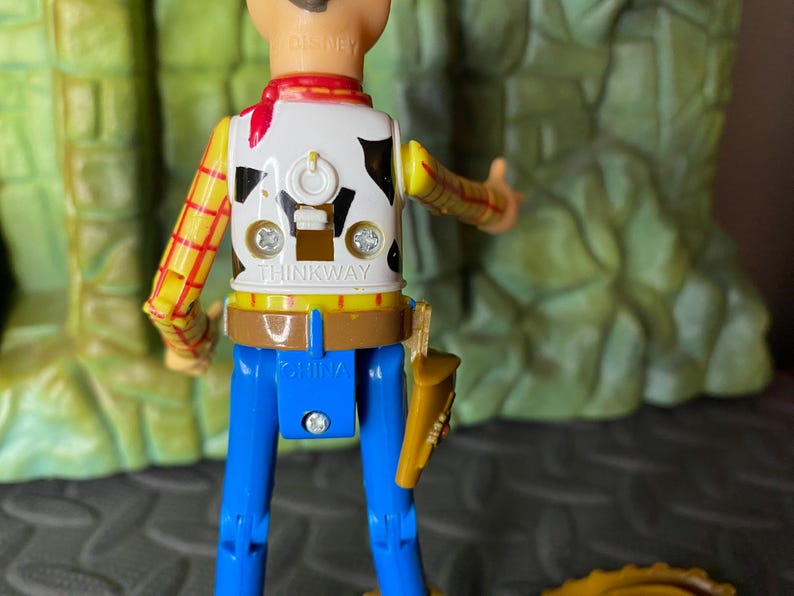 May include: A Woody action figure from the Toy Story franchise. The figure has a white vest with a black cowhide pattern, a yellow and red plaid shirt, blue pants, and a brown belt with a holster. The figure is marked "THINKWAY" and "CHINA".