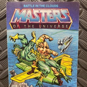 May include: A vintage Masters of the Universe trading card with a battle scene illustration. The title "Battle in the Clouds" is at the top. The artwork shows two muscular figures fighting, set against a blue sky with a futuristic vehicle. The card has text at the bottom.