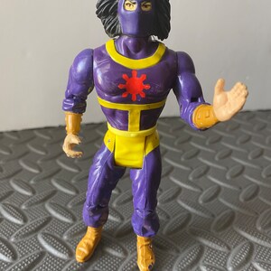Puede incluir: Figura de acción morada con una faja amarilla y un símbolo de sol rojo en el pecho. La figura tiene una máscara negra y cabello negro largo.