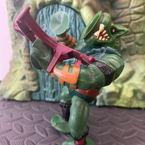 Puede incluir: Figura de acción verde de un personaje humanoide reptiliano, con detalles en naranja y rojo. La figura sostiene un arma roja y tiene un sombrero verde. El fondo presenta una estructura verde y marrón.