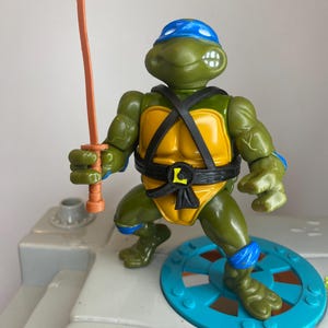 Può includere: Action figure di Leonardo, una delle Tartarughe Ninja, in posa da combattimento. La figura è verde con maschera blu, guscio giallo e cintura nera. Tiene una spada katana arancione. La figura è su una piattaforma azzurra.