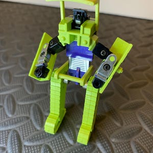 May include: A lime green and purple toy robot with a yellow canopy. The robot has black joints and silver accents. The toy is standing on a textured surface.