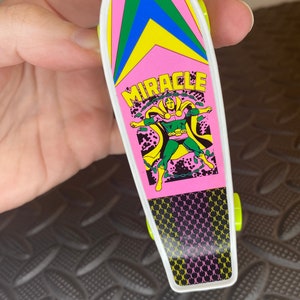 May include: A pink and green toy skateboard with a black and white graphic of a superhero with the word "MIRACLE" printed on the board.