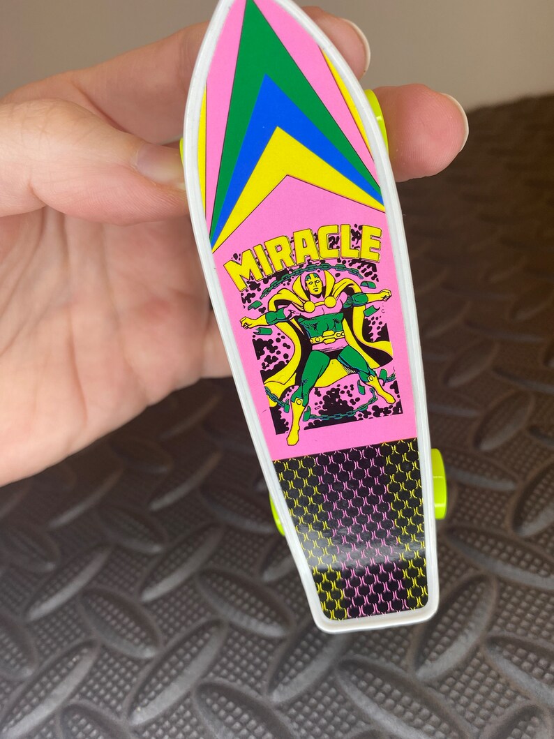 May include: A pink and green toy skateboard with a black and white graphic of a superhero with the word "MIRACLE" printed on the board.