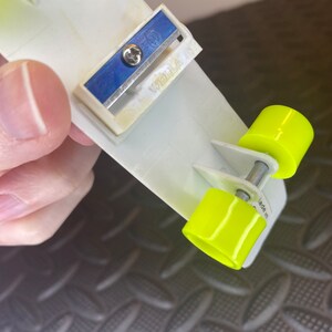 May include: A white plastic pencil sharpener with a blue blade and a yellow plastic base. The sharpener has two wheels on the bottom and the text "Made in" is visible on the side.