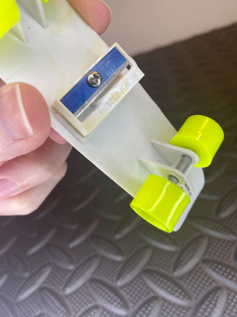 May include: A white plastic pencil sharpener with a blue blade and a yellow plastic base. The sharpener has two wheels on the bottom and the text "Made in" is visible on the side.