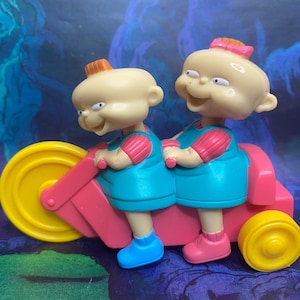 May include: A toy featuring two cartoon babies riding a pink and yellow tricycle. The babies have blonde hair, blue dresses, pink sleeves, and blue and pink shoes. The tricycle has a large yellow front wheel.