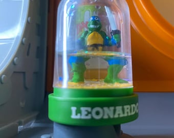 Puede incluir: Una figura de acción de Leonardo de las Tortugas Ninja Mutantes Adolescentes, verde y azul, en una cúpula de plástico transparente con una base de arena. La cúpula tiene la palabra "LEONARDO" impresa en la base.