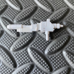 May include: White plastic toy gun with a detailed design. The gun has a barrel, a handle, and a trigger. It is a toy weapon for children.