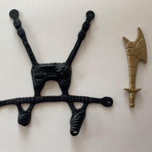 May include: Black plastic toy component with intricate detailing, alongside a gold-colored plastic toy axe. The black piece has a unique, symmetrical design with multiple arms and circular ends. The axe features a detailed blade.