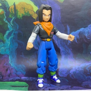 May include: A plastic action figure of a character with black hair, wearing a dark gray short-sleeved shirt over a white long-sleeved shirt, an orange neck scarf, blue pants with a brown belt, green socks, and blue and white sneakers.