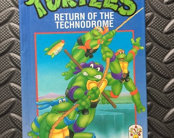 May include: A blue and green Teenage Mutant Hero Turtles book cover featuring the four turtles hanging from a rope. The book title is "Return of the Technodrome".