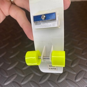 Puede incluir: Un sacapuntas de pl&aacute;stico blanco con forma de patineta con ruedas amarillas. El sacapuntas tiene una cuchilla azul y dice "Wella" en el lateral. Dice "Hecho en China" en la parte inferior.