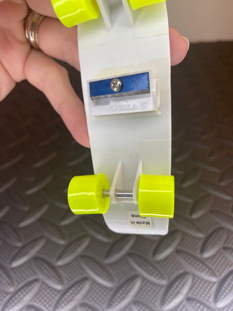 Puede incluir: Un sacapuntas de pl&aacute;stico blanco con forma de patineta con ruedas amarillas. El sacapuntas tiene una cuchilla azul y dice "Wella" en el lateral. Dice "Hecho en China" en la parte inferior.