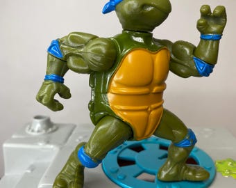 Pode incluir: Figura de ação das Tartarugas Ninja em pose de combate. A figura é principalmente verde com uma bandana azul, concha laranja e detalhes azuis. Está sobre uma plataforma cinza com uma base circular azul e verde.