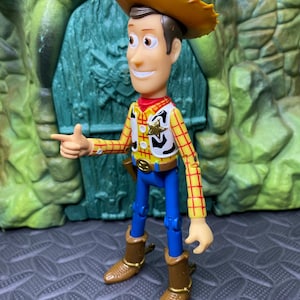 May include: A Woody action figure, featuring a yellow plaid shirt, blue pants, and a brown cowboy hat and boots. The figure has a sheriff's badge and is pointing with one hand.