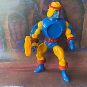 Puede incluir: Una figura de acción azul y amarilla con un casco amarillo y una franja roja. La figura sostiene un disco amarillo en su mano derecha. La figura está de pie sobre una superficie marrón y morada.