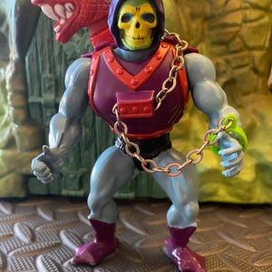 May include: A green and purple Skeletor action figure with a red dragon head on his back. He is wearing purple and red armour with a chain attached to his chest. The figure is standing on a grey and white patterned surface.