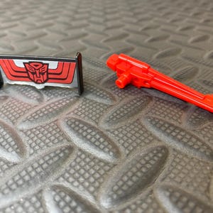May include: A red and black toy accessory, featuring a red emblem with a robot face, and a red toy gun. The objects are on a textured gray surface. The toy accessories are likely for a robot action figure.