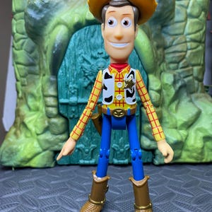 May include: A Woody action figure, featuring a yellow cowboy hat, a red and yellow plaid shirt, a white vest with black spots, blue pants, and brown boots. The figure has a sheriff's badge and a belt buckle. The background is a green and gray structure.