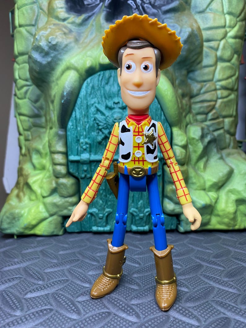 May include: A Woody action figure, featuring a yellow cowboy hat, a red and yellow plaid shirt, a white vest with black spots, blue pants, and brown boots. The figure has a sheriff's badge and a belt buckle. The background is a green and gray structure.