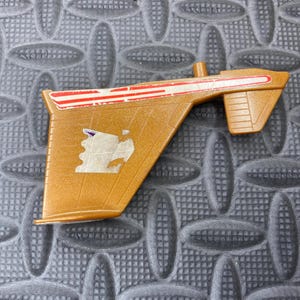 May include: A gold-coloured toy ray gun with red and white stripes on the top. The toy has a stylized design with a small emblem on the side. The ray gun is set against a textured grey background.