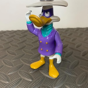 Puede incluir: Una figura de acción de Darkwing Duck que lleva un sombrero gris de ala ancha, una chaqueta morada de doble botonadura con botones amarillos, un pañuelo turquesa y guantes blancos. El personaje tiene pico amarillo, pies amarillos y antifaz morado.