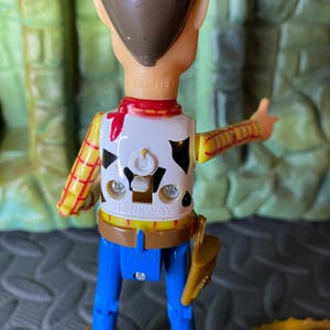 May include: A Woody action figure from the Disney Pixar Toy Story franchise. The figure is wearing a white vest with black cow spots, a yellow shirt with red stripes, blue pants, and brown boots. The figure has a brown cowboy hat.