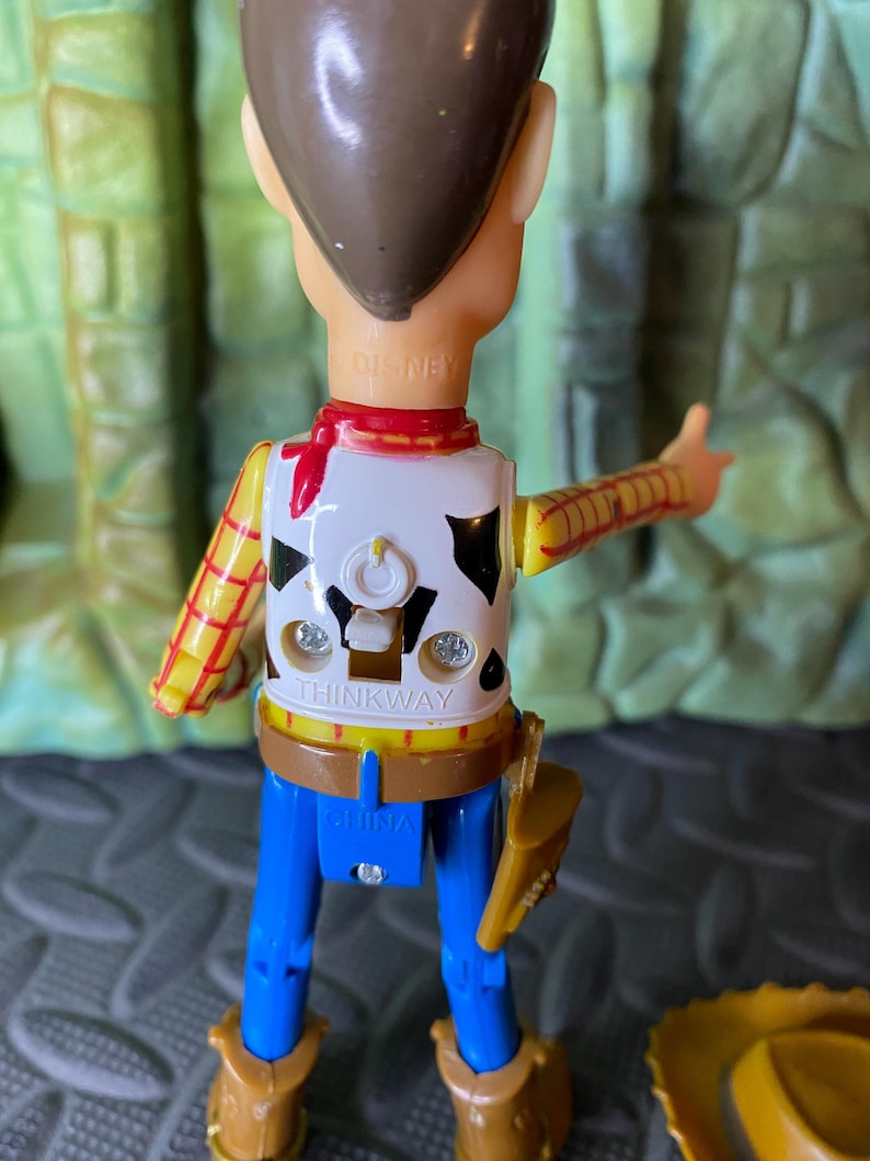 May include: A Woody action figure from the Disney Pixar Toy Story franchise. The figure is wearing a white vest with black cow spots, a yellow shirt with red stripes, blue pants, and brown boots. The figure has a brown cowboy hat.