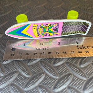 May include: A pink and white toy skateboard with a green, yellow, and blue design. The skateboard has a character with a green costume and a yellow mask on it. The text "MIRACLE" is printed on the skateboard. The skateboard is about 4 inches long.