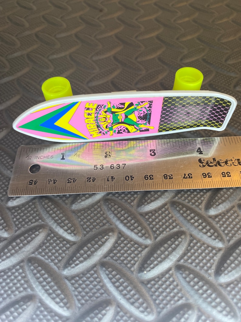 May include: A pink and white toy skateboard with a green, yellow, and blue design. The skateboard has a character with a green costume and a yellow mask on it. The text "MIRACLE" is printed on the skateboard. The skateboard is about 4 inches long.