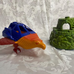May include: A blue, orange, and red plastic toy spaceship with a bird-like head and wings. The spaceship is sitting on a white surface next to a green plastic toy castle.